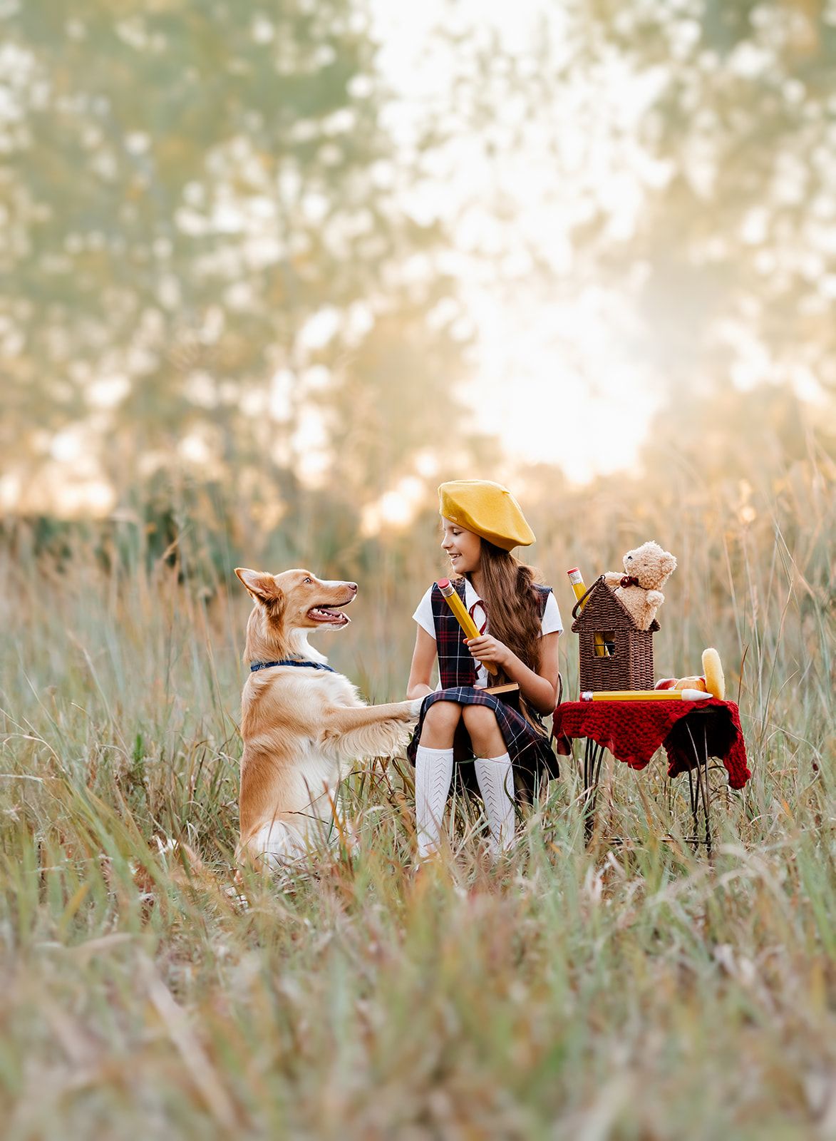 Young girl in yellow beret enjoys outdoor tea party with golden dog and teddy bear in sunlit meadow.