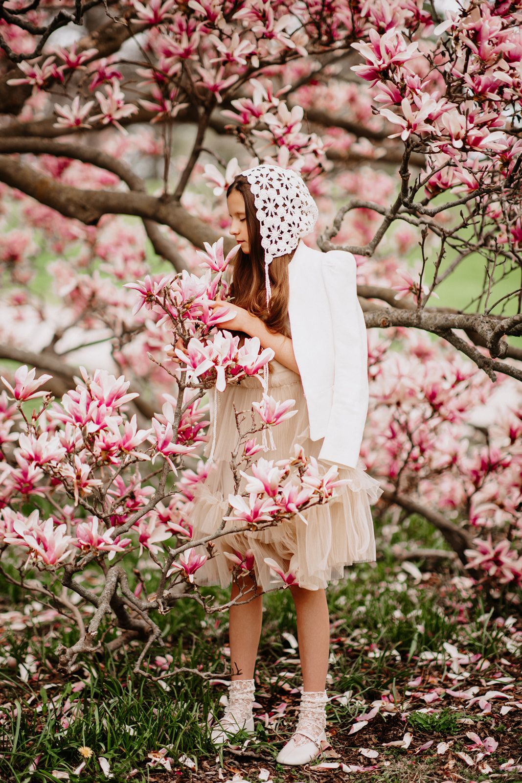 Young girl in beige dress and white lace bonnet smelling pink magnolia blossoms in a flowering spring garden.