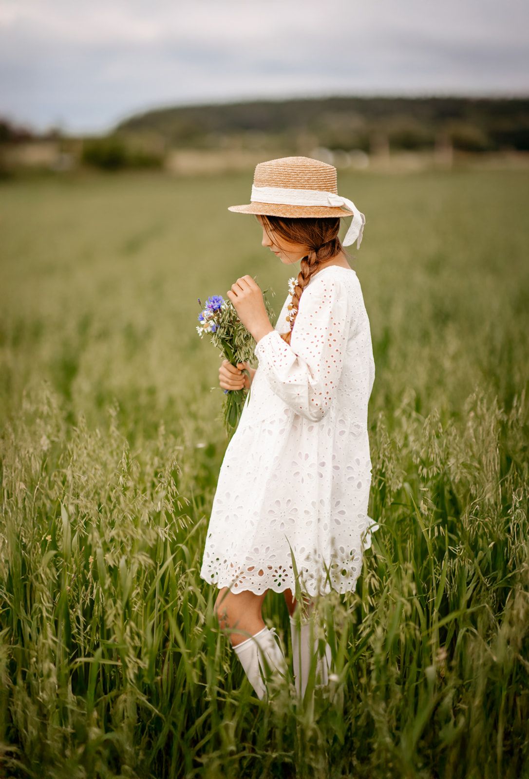 A young girl in a white lace dress and straw hat holds wildflowers in a lush green field.