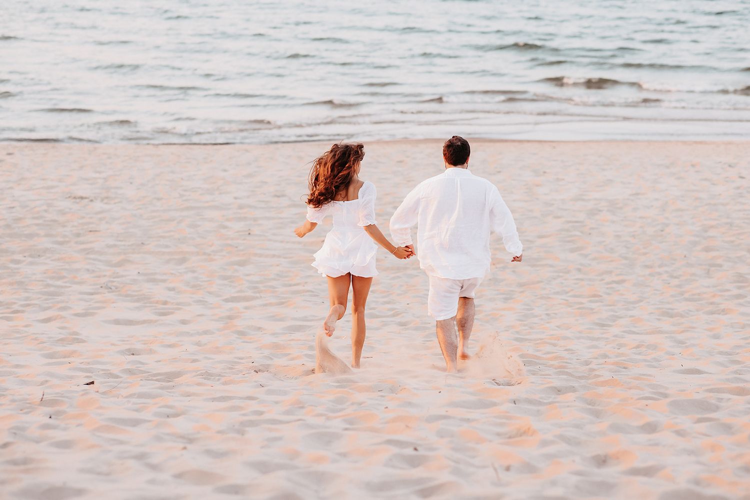 Two children in white outfits holding hands and running on a sandy beach near the ocean at sunset.