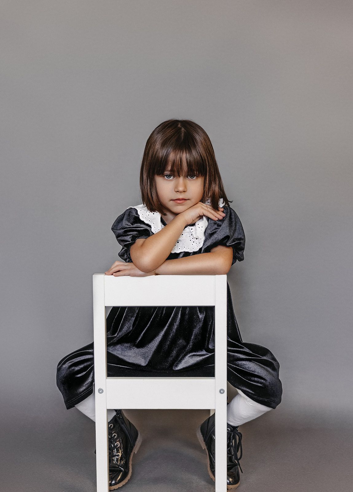Young girl in black velvet dress with white collar sits on white chair, arms crossed, gray background.