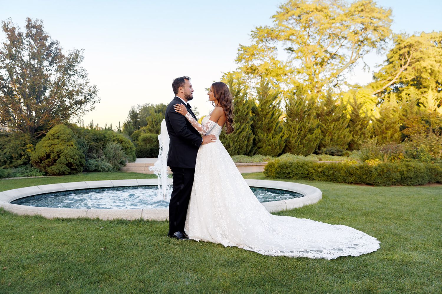 Bride and groom share a romantic moment by a garden fountain at dusk in elegant wedding attire.