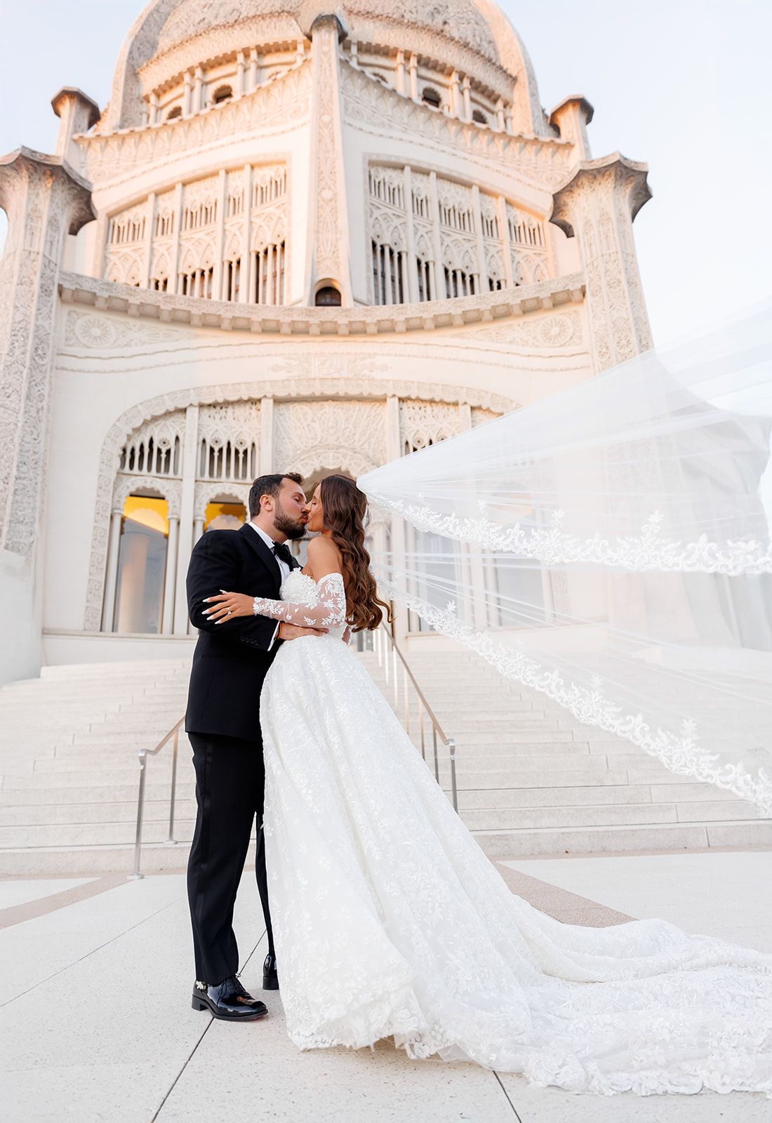 Bride and groom share a kiss on steps before ornate white Bahai Temple, veil flowing dramatically in the wind.