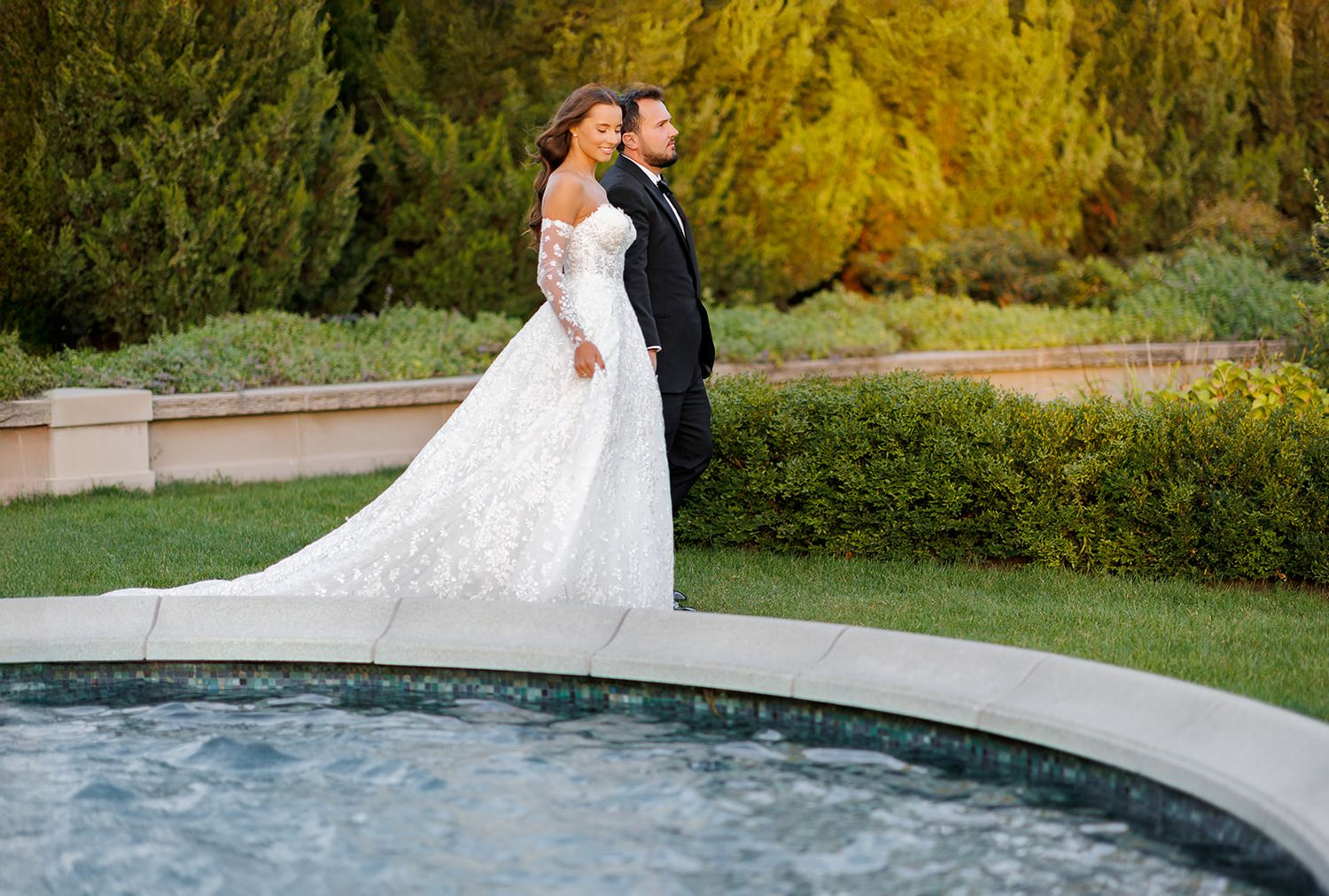 Elegant bride and groom stand by garden pool, bride wearing long-sleeve lace wedding gown with flowing train.