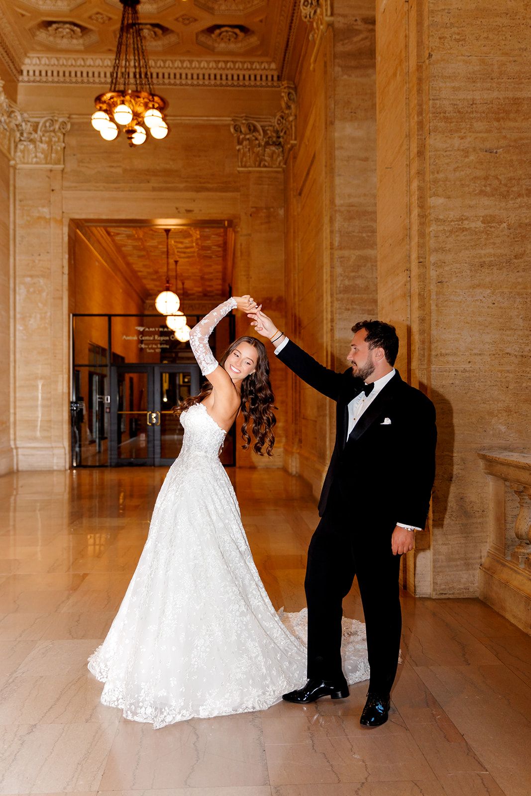 Bride in white lace gown spins under groom's hand in black tuxedo inside elegant marble hall with chandelier.