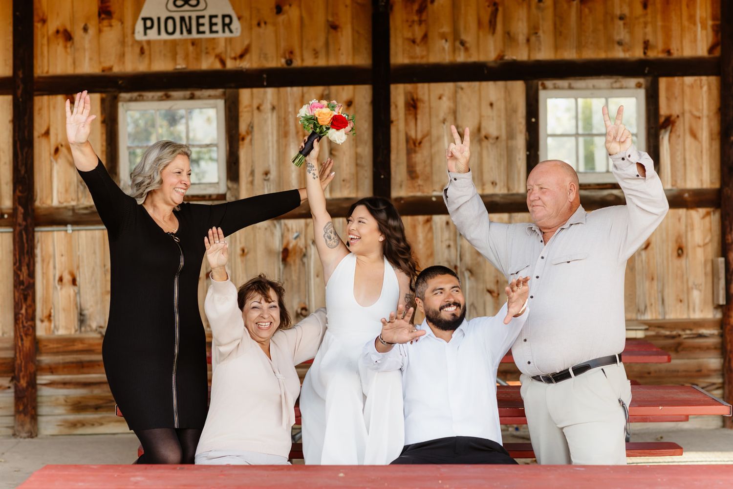 Joyful wedding celebration in a rustic barn with bride, groom, and family raising arms in excitement.