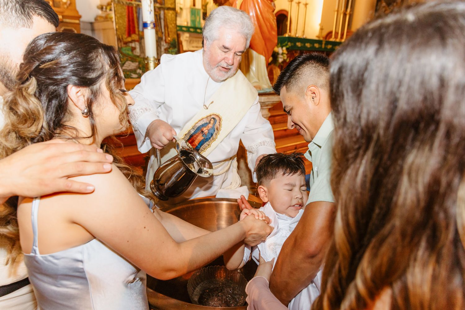 Priest pours holy water over baby during baptism with family gathered around font.