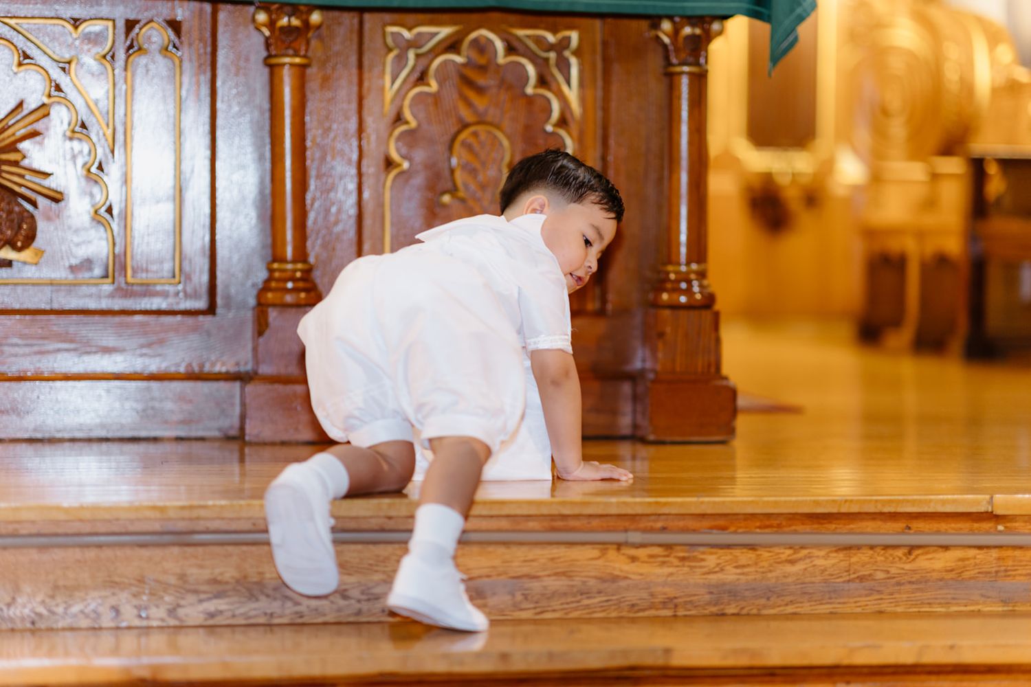 Young toddler in white outfit crawling on wooden church floor during baptism ceremony.