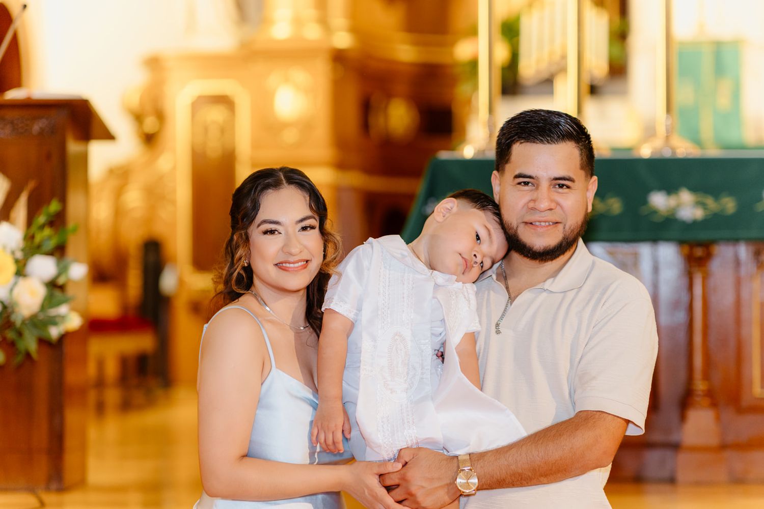 A happy family of three poses with their baby in white baptism outfit inside a decorated church.