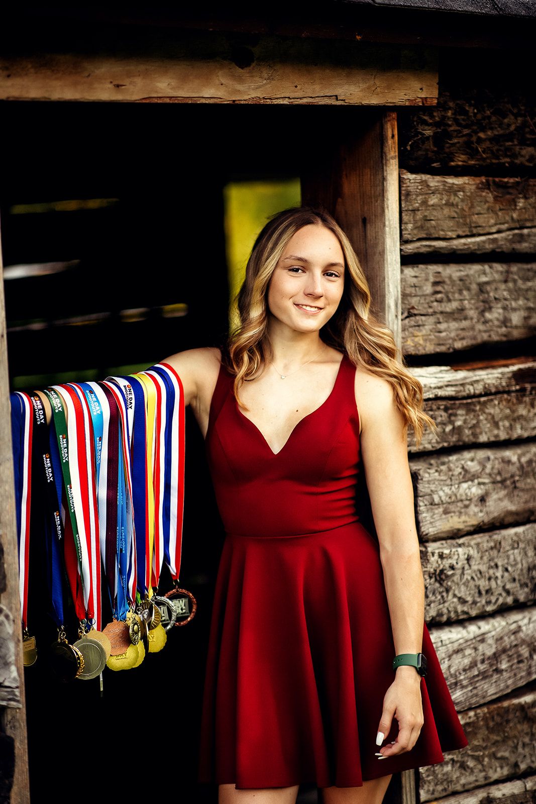 Young woman in red dress displays a collection of colorful medals hanging in a rustic wooden doorway.