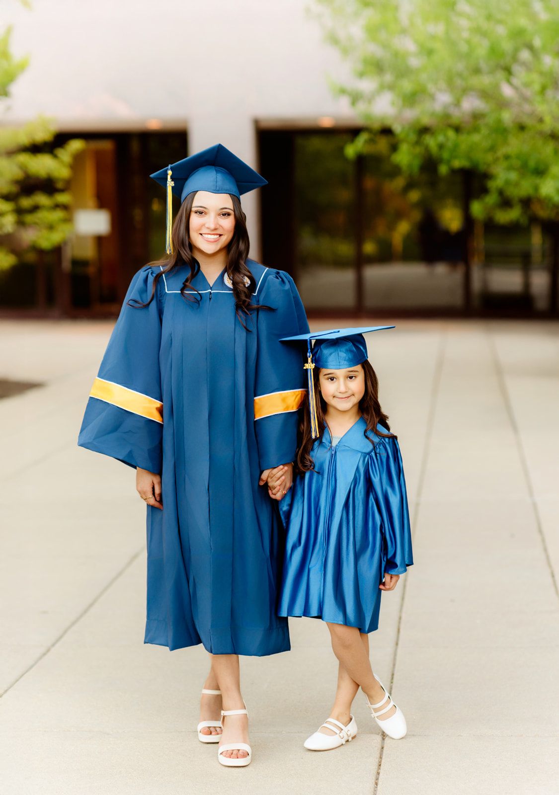 Older student and young girl both wearing matching navy blue graduation caps and gowns outdoors.