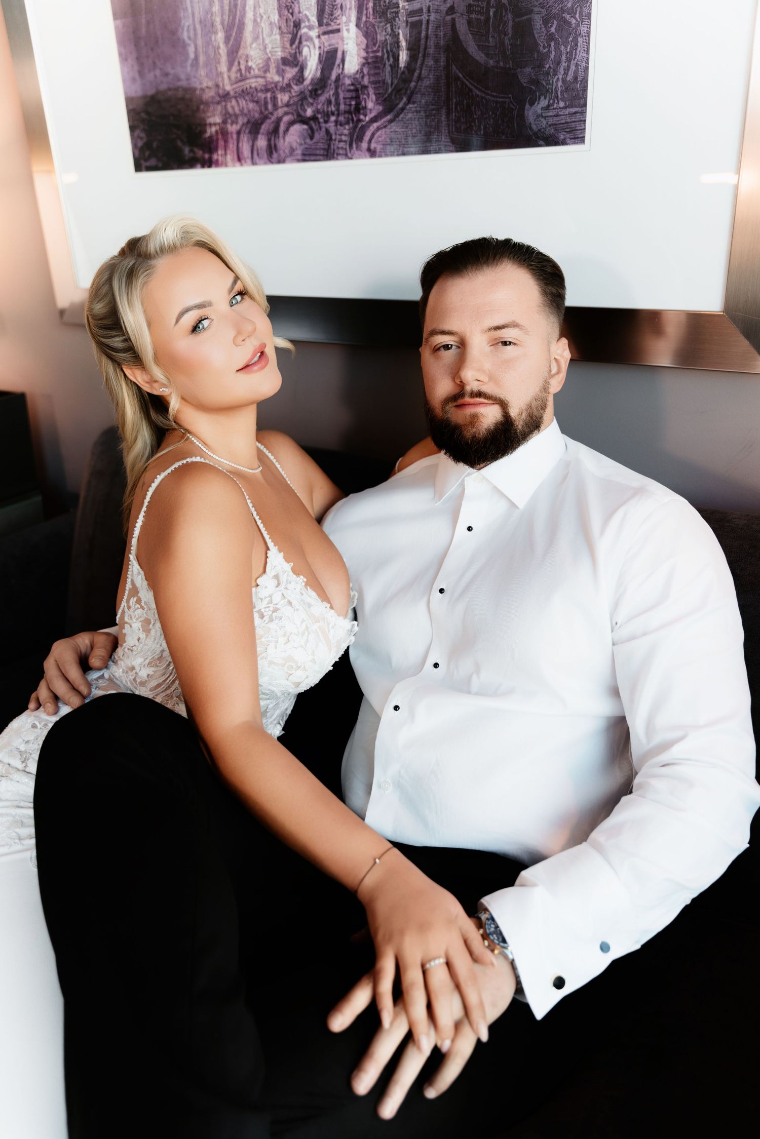 Bride in white lace dress and bearded groom in white shirt pose together on sofa.