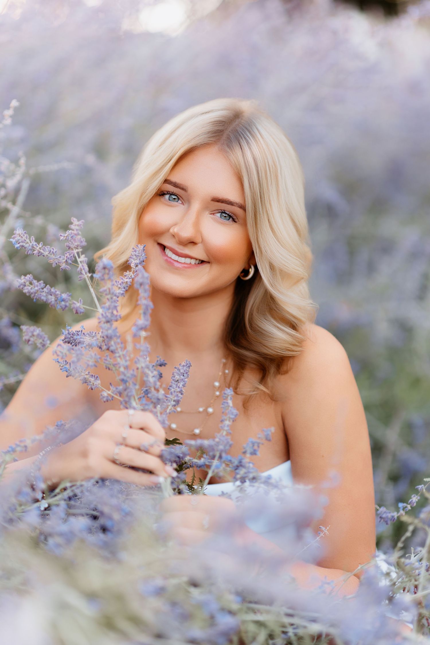 Blue-eyed blonde woman smiling while holding lavender sprigs in a purple flower field.
