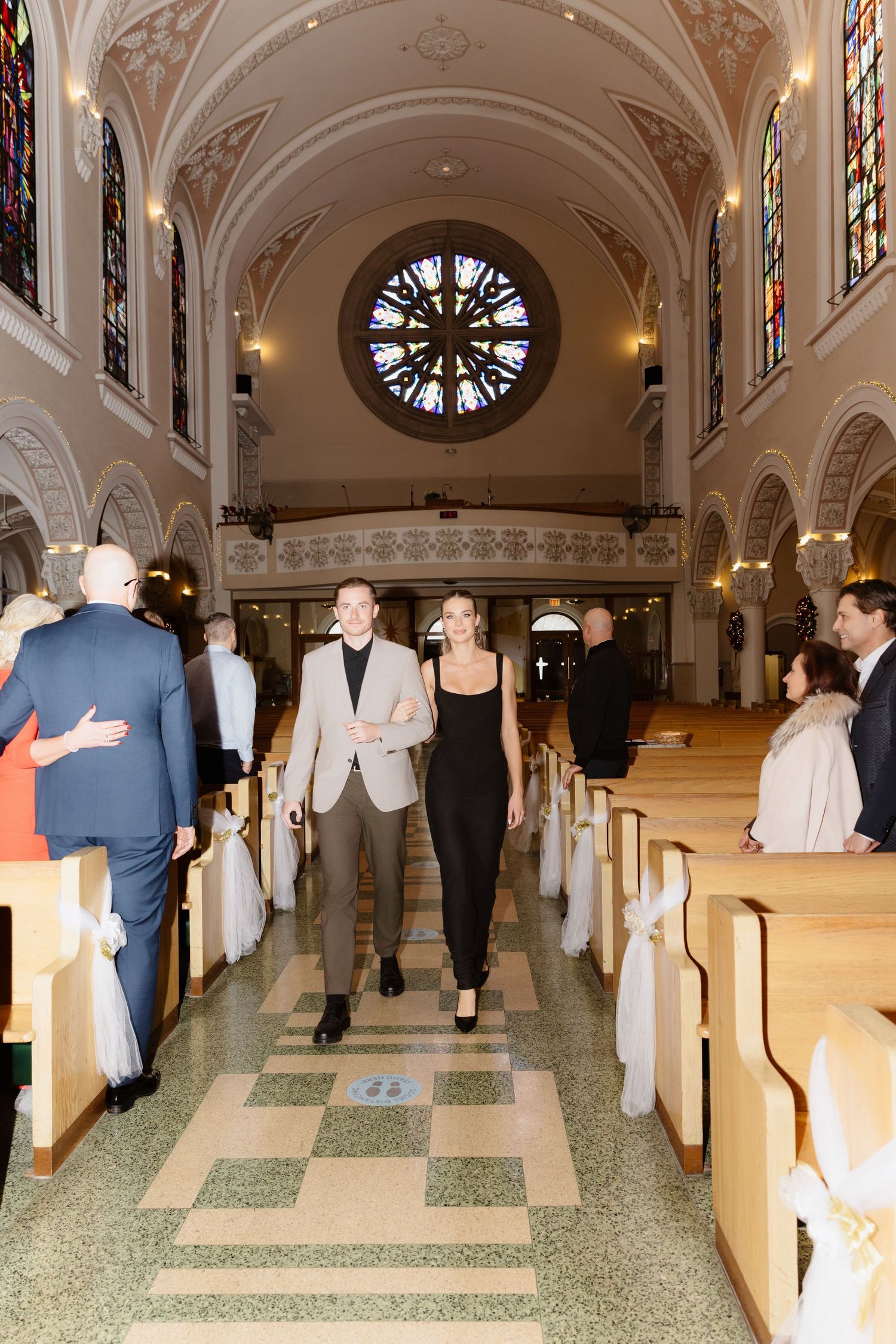 A couple walks down the aisle of a beautiful church with stained glass windows and wooden pews.