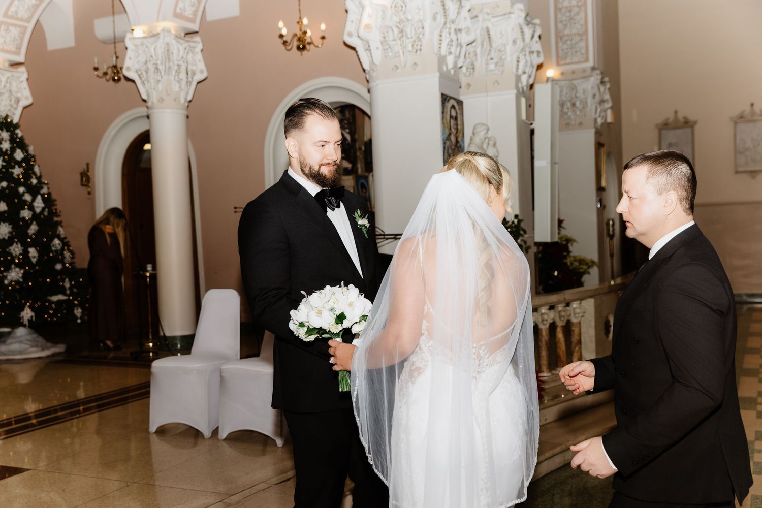 Groom in tuxedo smiles at bride in lace wedding dress holding white bouquet inside festive church.