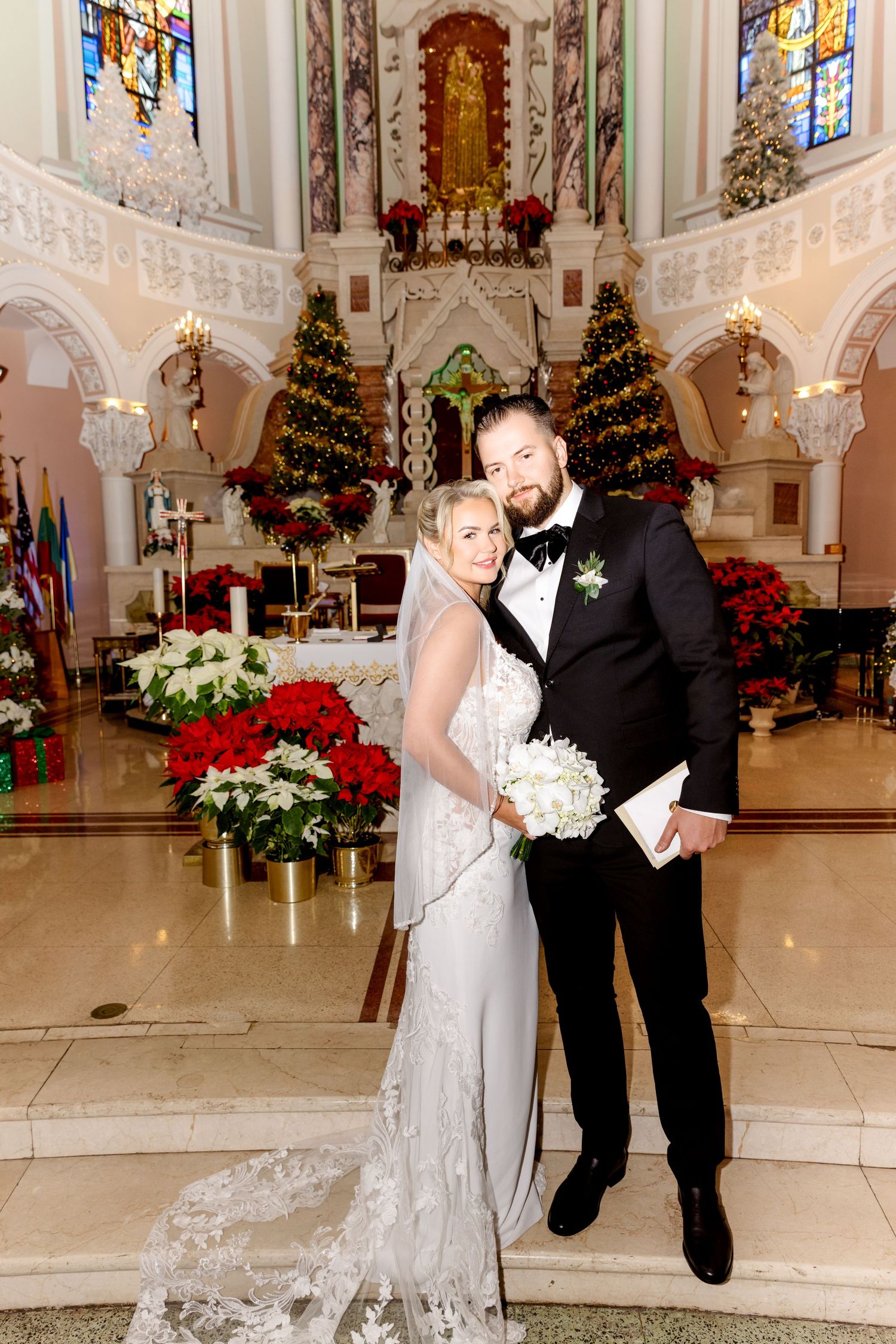 Bride and groom pose elegantly at church altar surrounded by Christmas trees and red poinsettia floral arrangements.