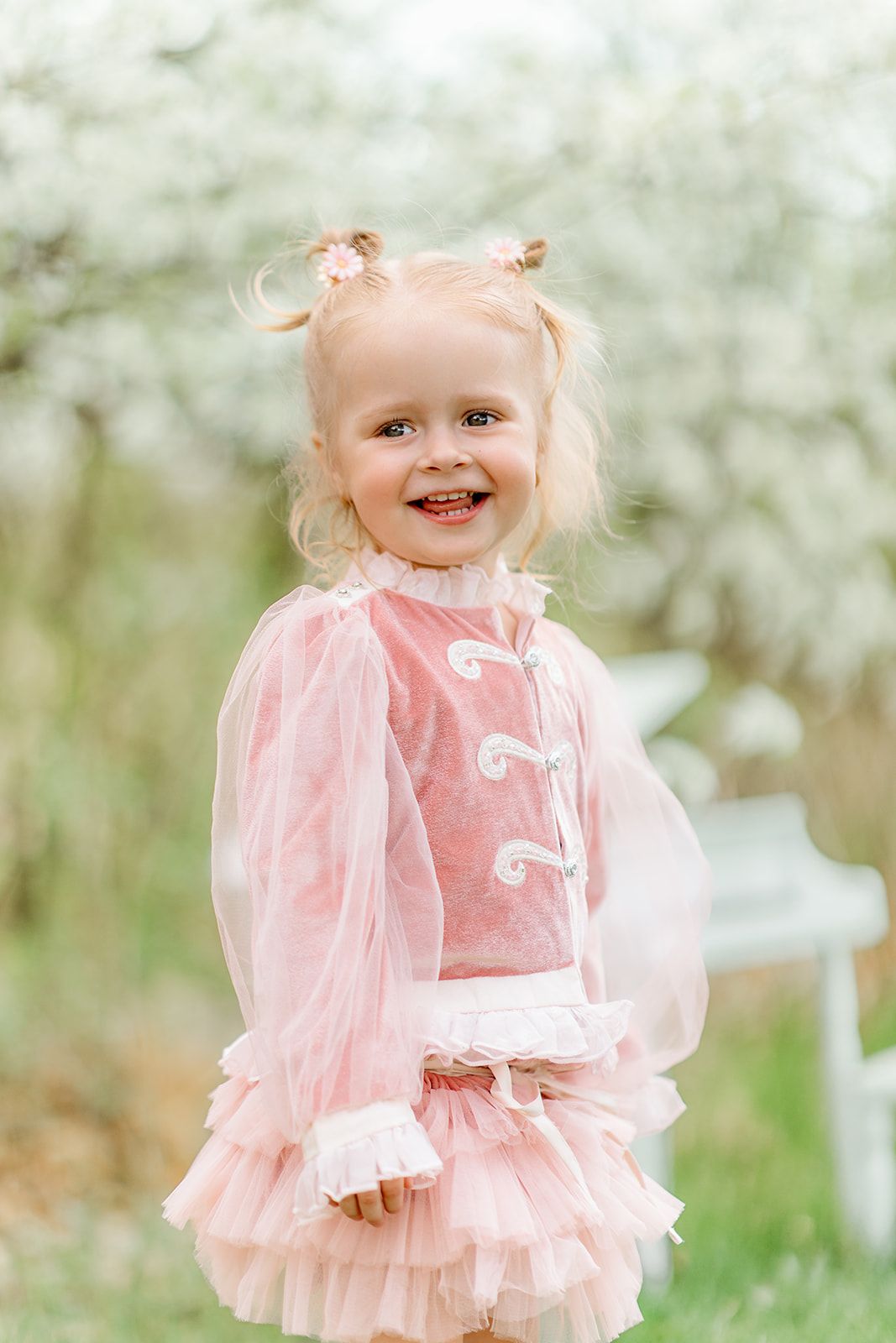 A cheerful toddler girl in a pink tulle dress smiles outdoors among blooming white trees in spring.
