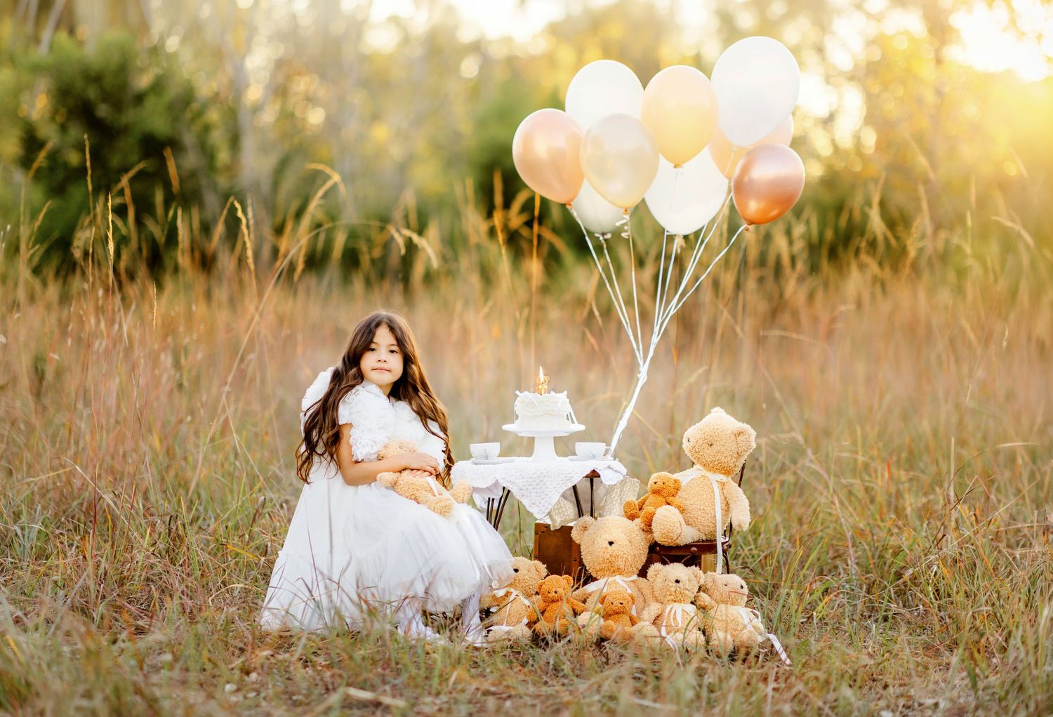 Girl in white dress sits in field with teddy bears, balloons, and cake for an outdoor birthday celebration.