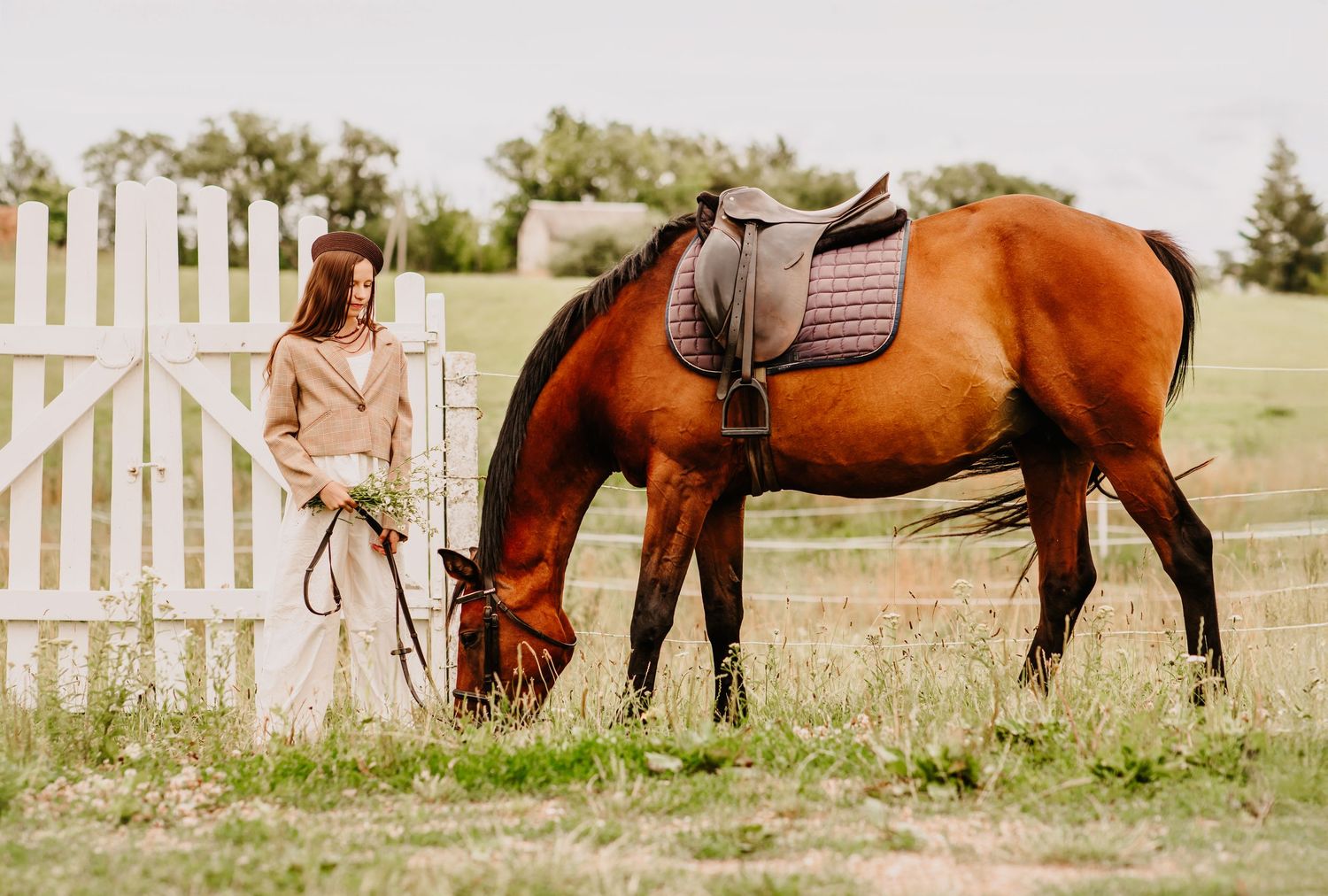 Woman in beige coat stands by white fence as a saddled bay horse grazes in green pasture.
