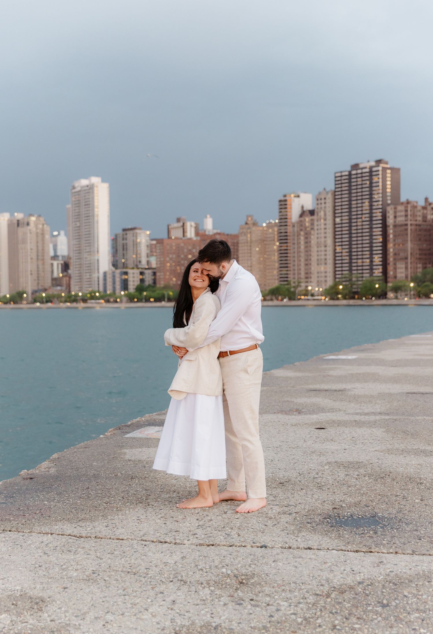 Man kissing womans head while embracing on Chicago lakefront pier with city skyline.