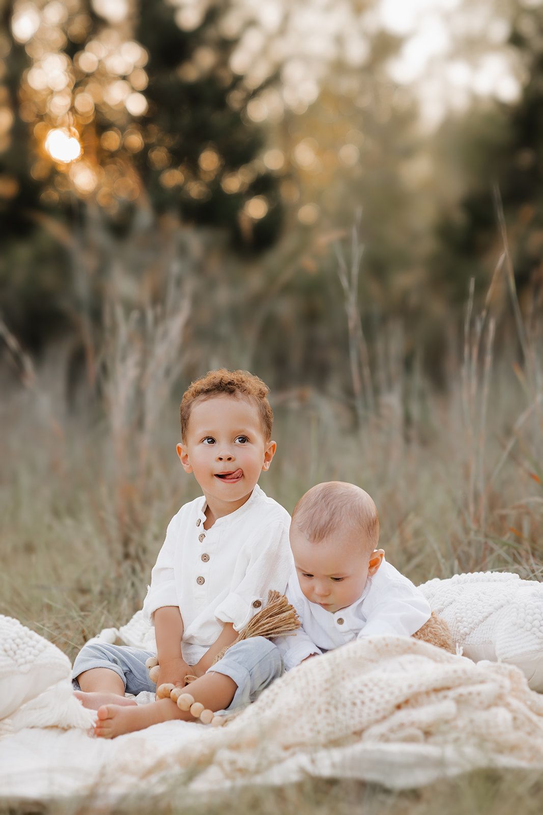 Two young brothers in white shirts sitting on a blanket in a sunlit meadow at golden hour.