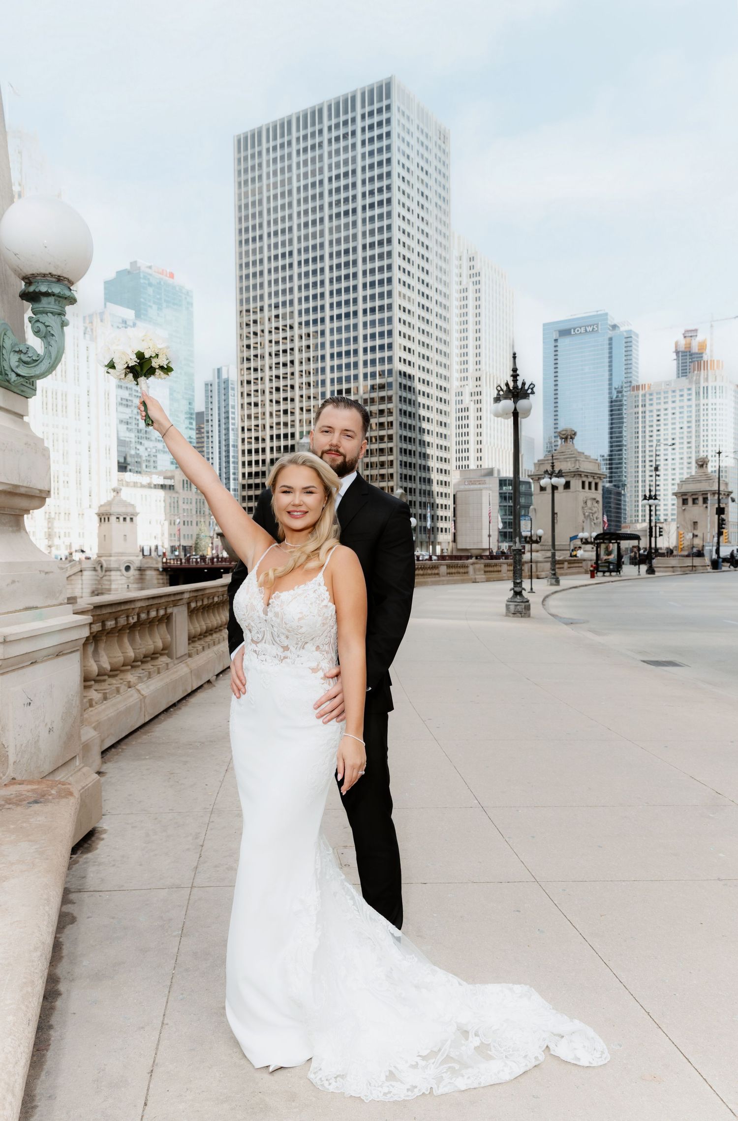 Joyful bride raising bouquet with groom on a Chicago bridge, skyscrapers visible in background.
