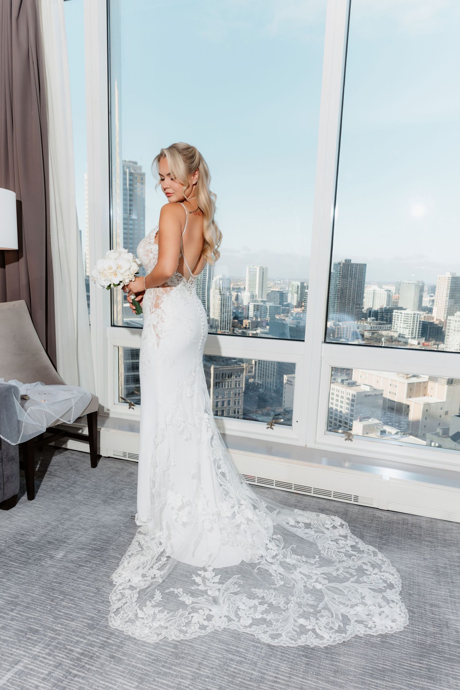 Bride in backless lace gown with cathedral train looks over shoulder holding bouquet before city windows.