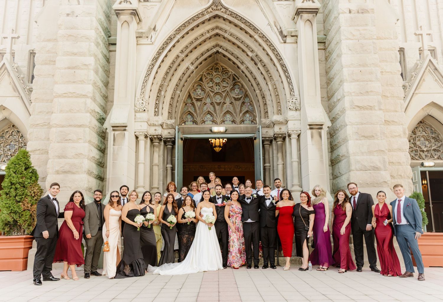 Large wedding party poses together outside ornate Gothic church entrance.