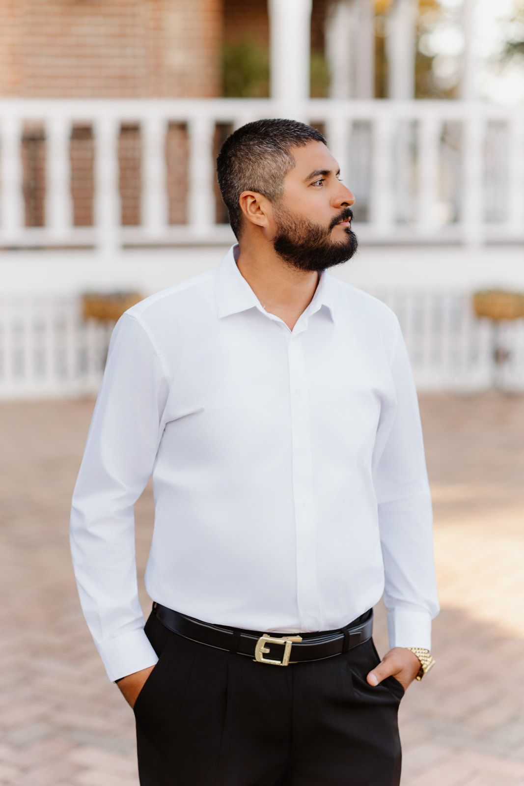 Bearded groom in white dress shirt and black pants standing outdoors near brick building.