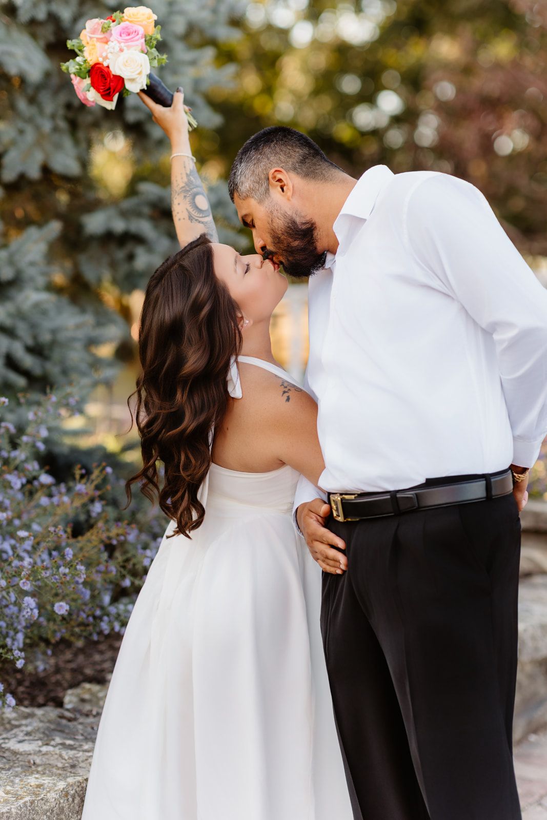 Bride and groom share romantic kiss outdoors, bride raises colorful bouquet while wearing white halter dress.