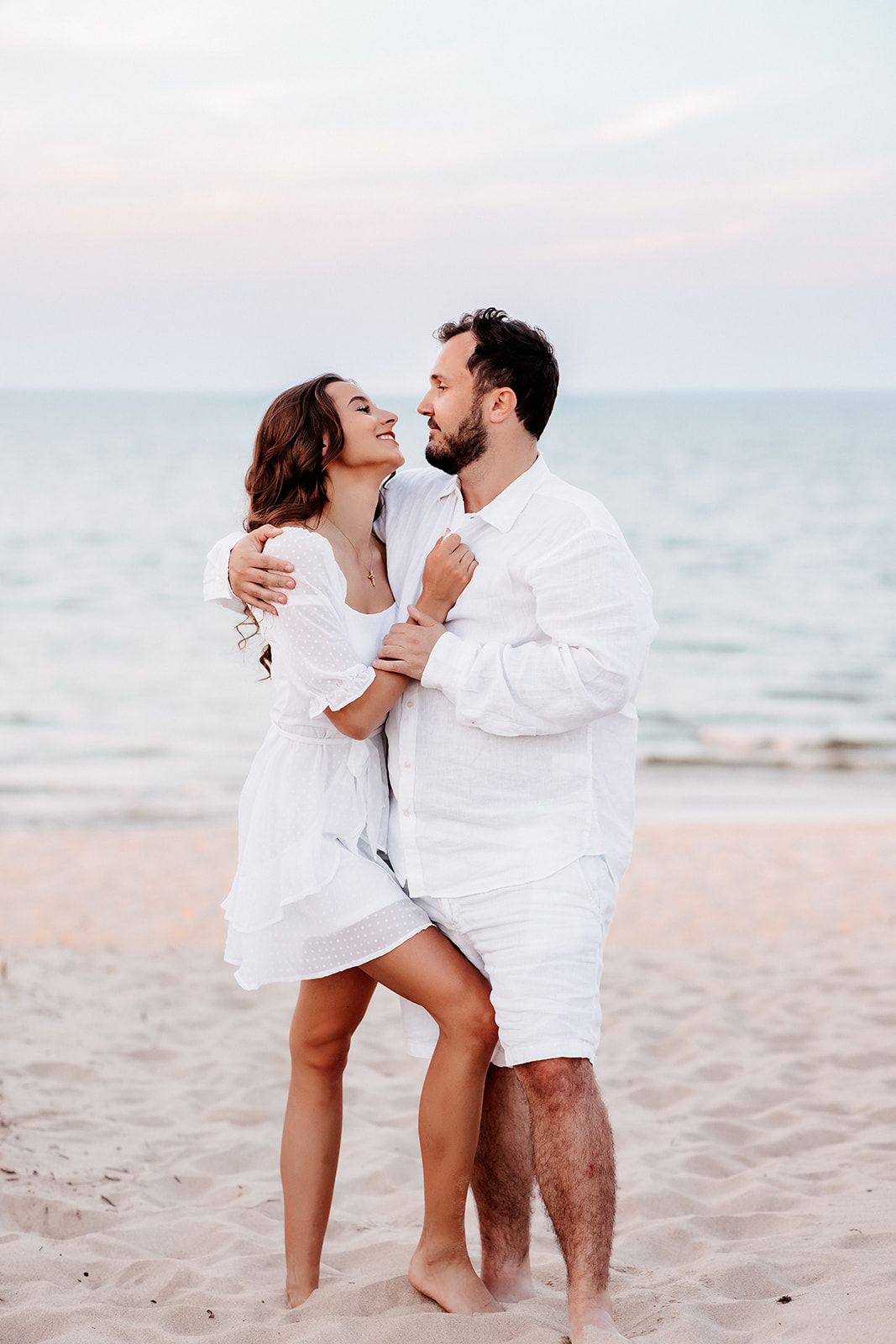 A romantic couple in matching white outfits embrace and smile on a sandy beach at sunset.