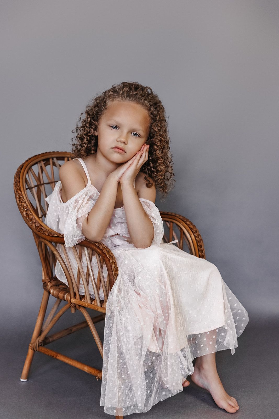 Young curly-haired girl in white polka dot dress sitting in wicker chair with sad expression.