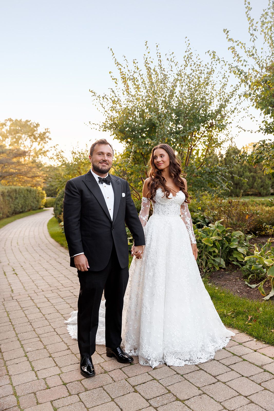 A bride in a lace gown and groom in a tuxedo hold hands on a garden pathway during their wedding day.