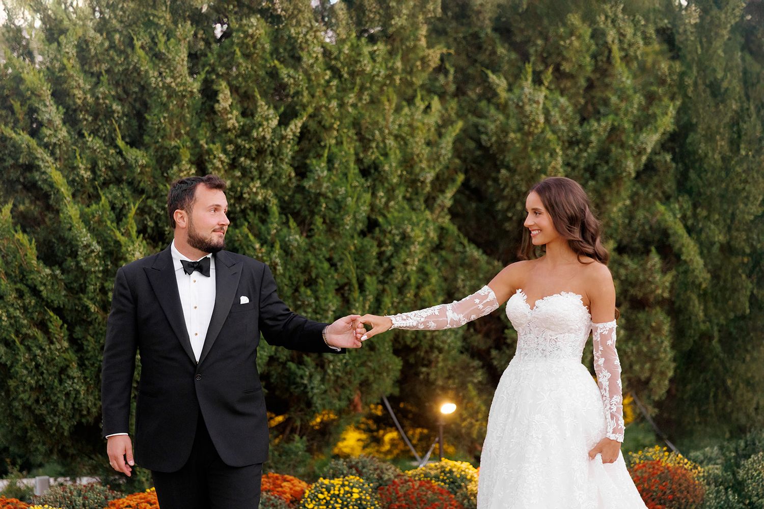 Bride and groom hold hands outdoors among lush green trees and colorful autumn flowers during wedding portraits.