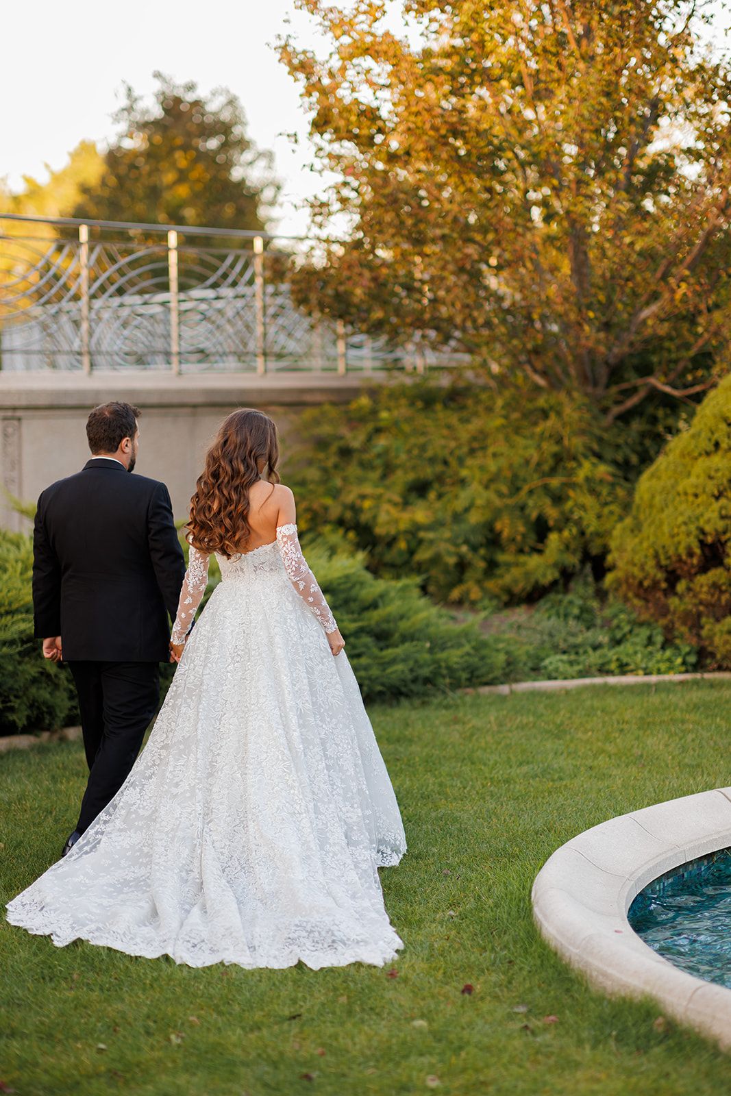 Bride in lace gown and groom in black tuxedo walk hand-in-hand through a lush garden near a fountain.