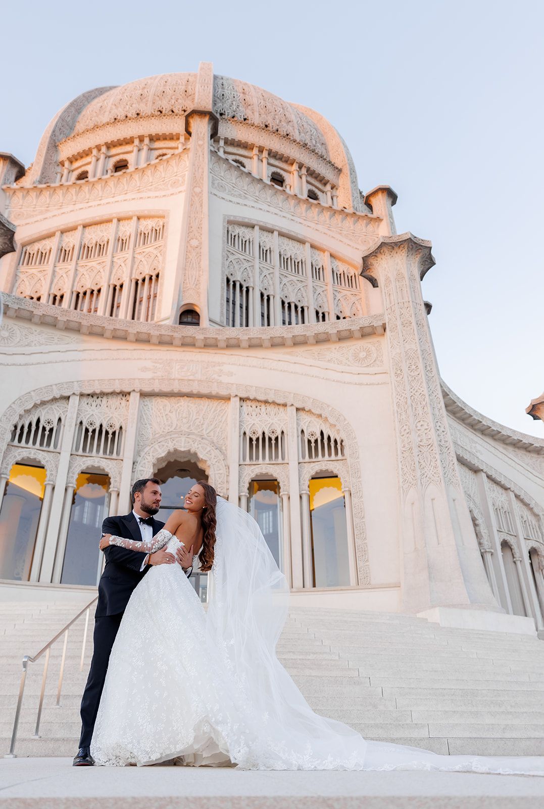 Bride and groom embrace on steps of ornate Bahai Temple with domed architecture glowing at sunset.