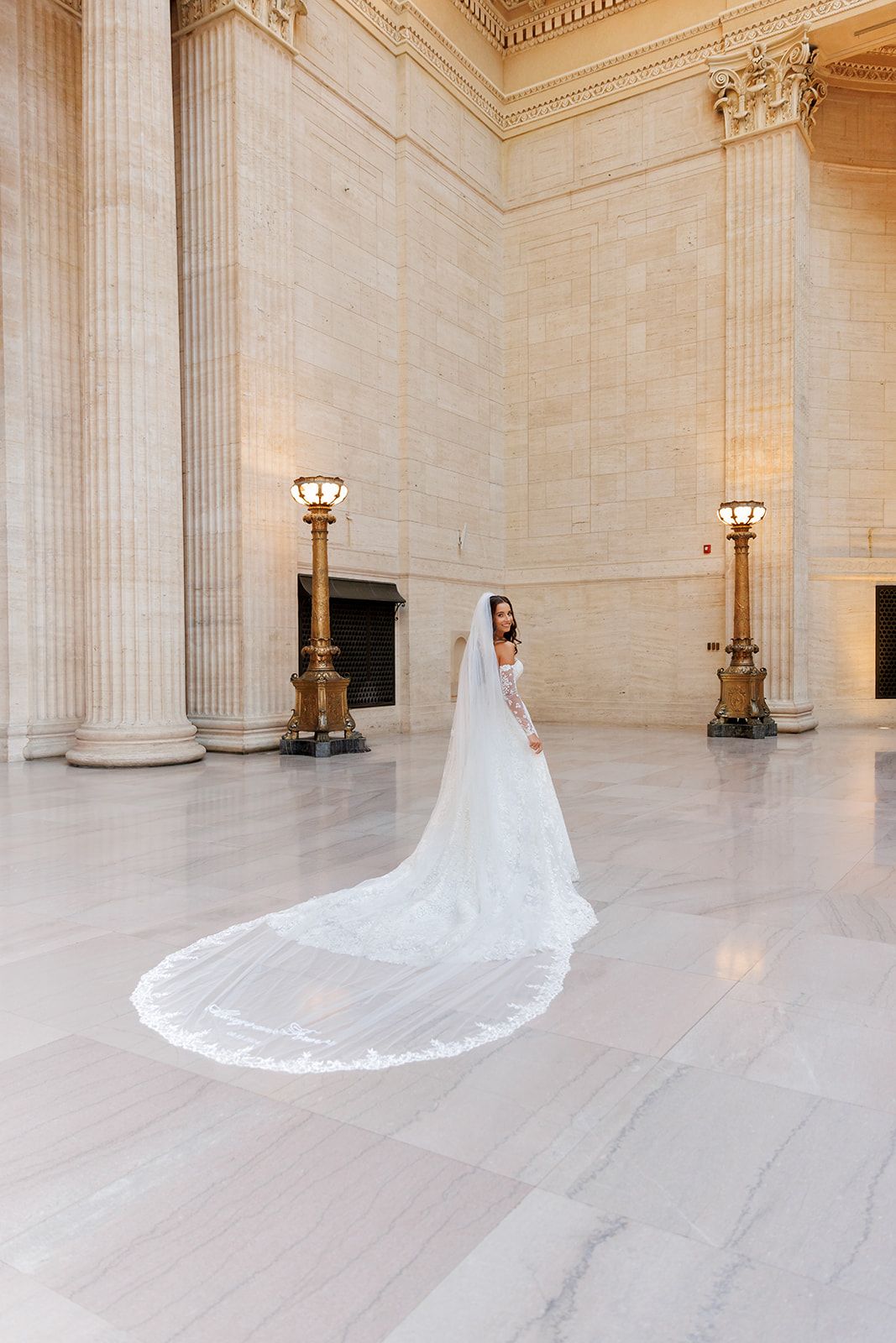 Bride in a white wedding gown with a long cathedral veil stands in a grand marble hall with tall columns.