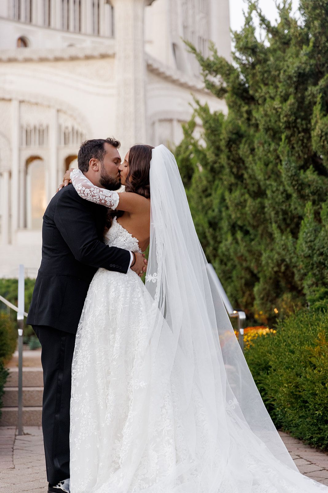 A bride and groom share a romantic kiss outside a grand white building, with the bride wearing a lace gown and long veil.