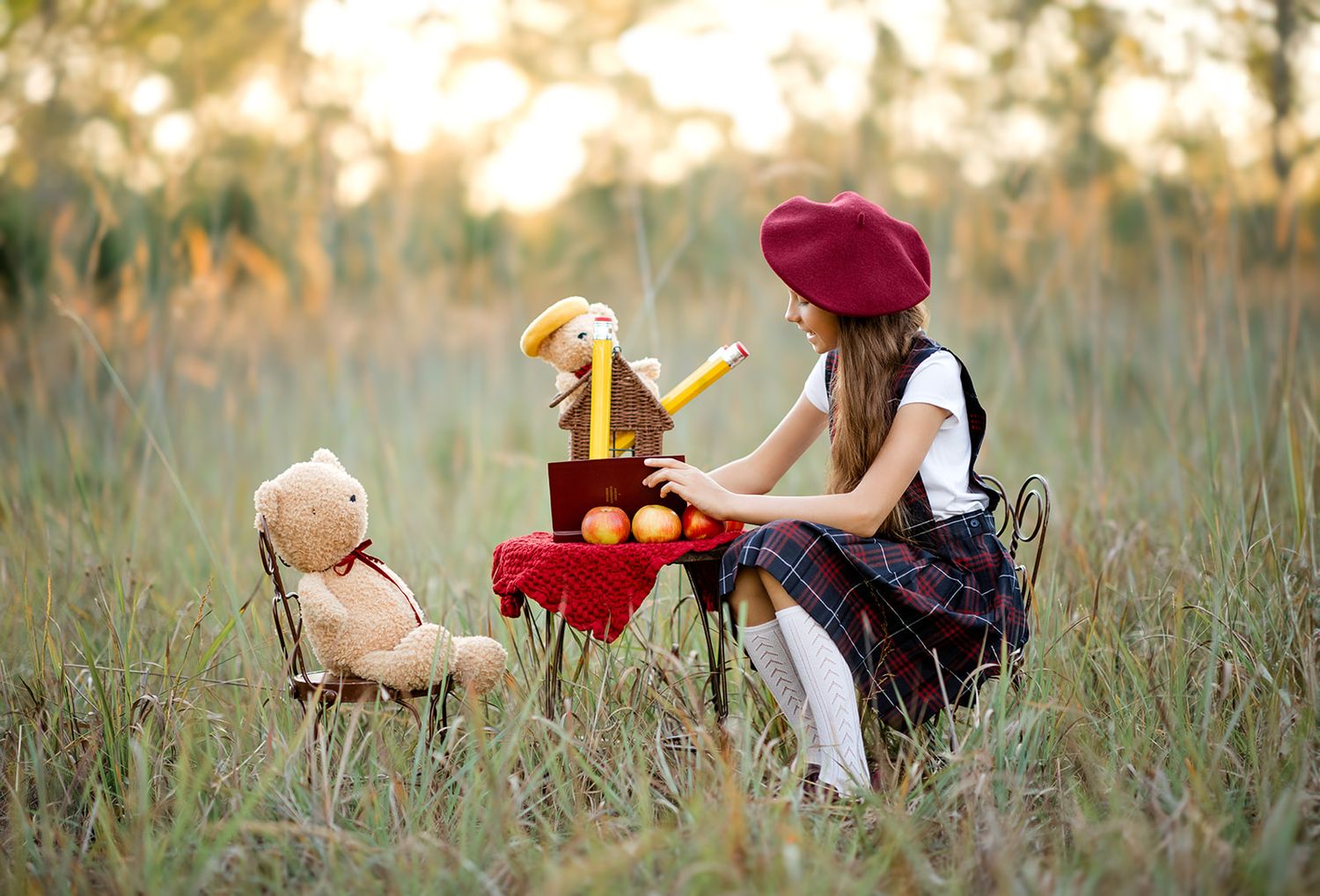 A young girl in a red beret hosts a tea party with stuffed animals in a grassy field.