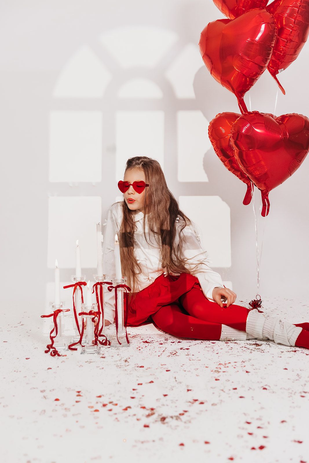 Young woman in red pants and heart sunglasses sits on floor with red heart balloons and candles for Valentine's Day.