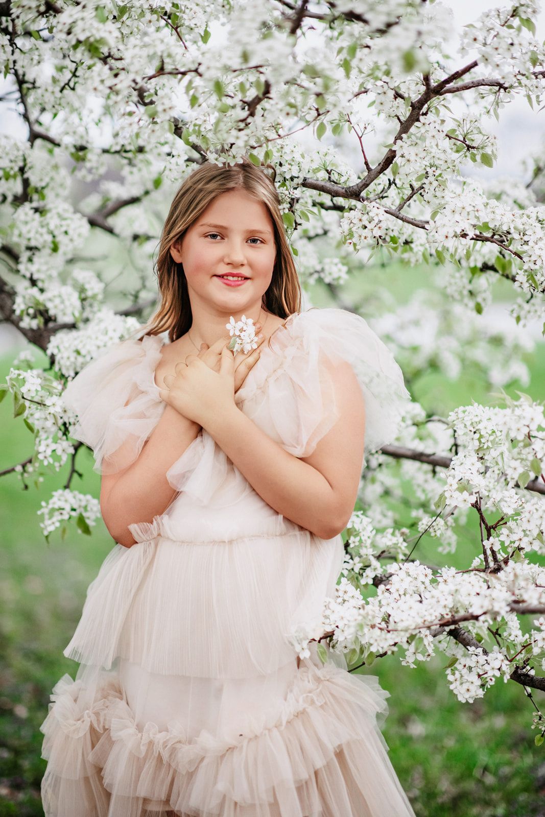 Young girl in a blush tulle dress holding white blossoms, smiling among flowering tree branches outdoors.