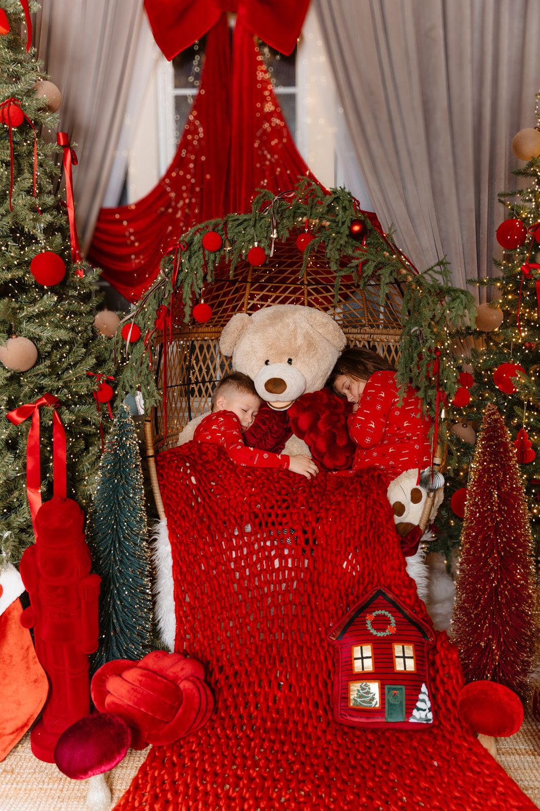 Child sleeping with teddy bear in festive Christmas cradle surrounded by decorated trees and red holiday decor.