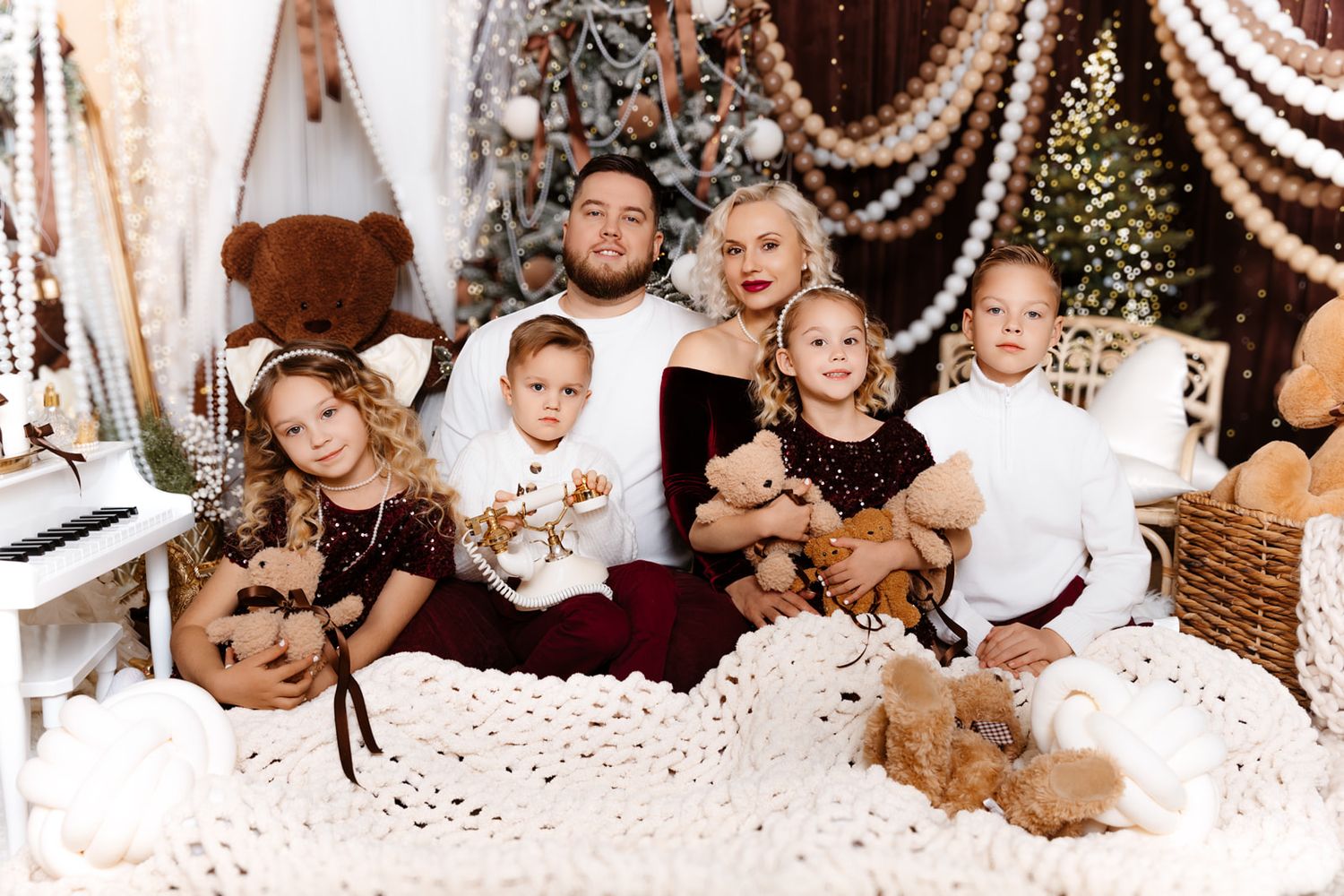 Family of six poses for a festive Christmas photo with holiday decorations, teddy bears, and a decorated tree in background.