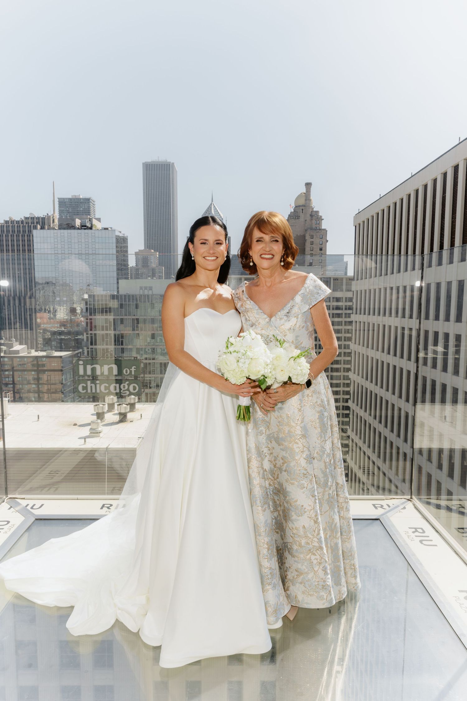 Bride in strapless white ball gown poses with mother on rooftop overlooking Chicago skyline.