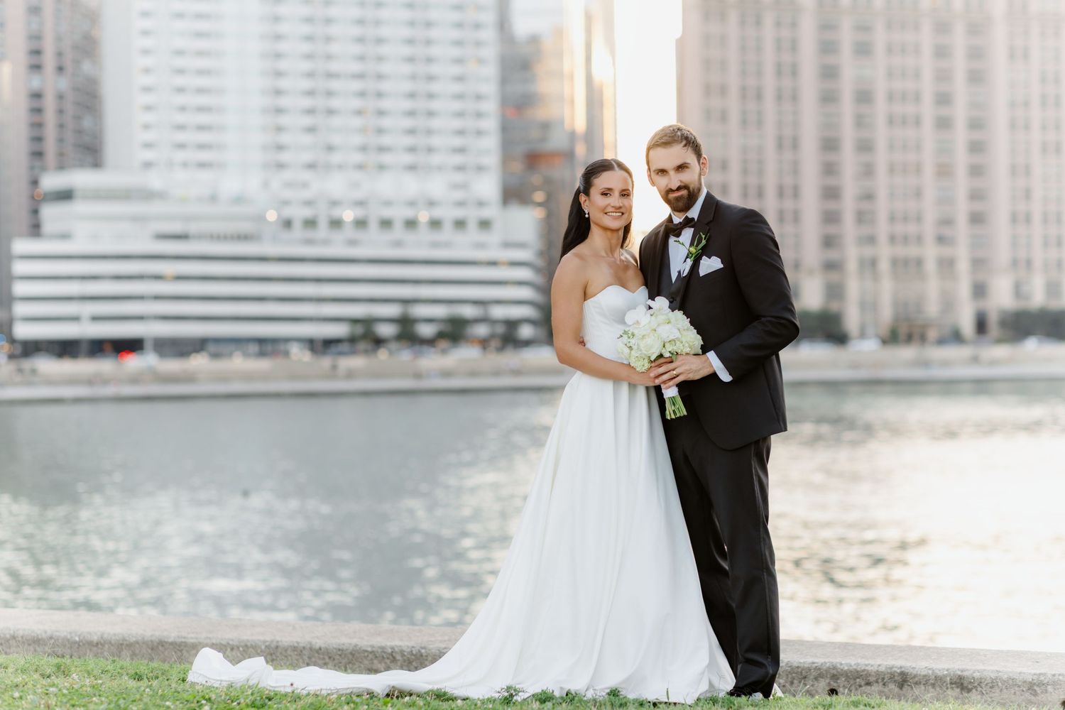 Elegant bride and groom pose together holding white bouquet by the waterfront with city skyline behind them.