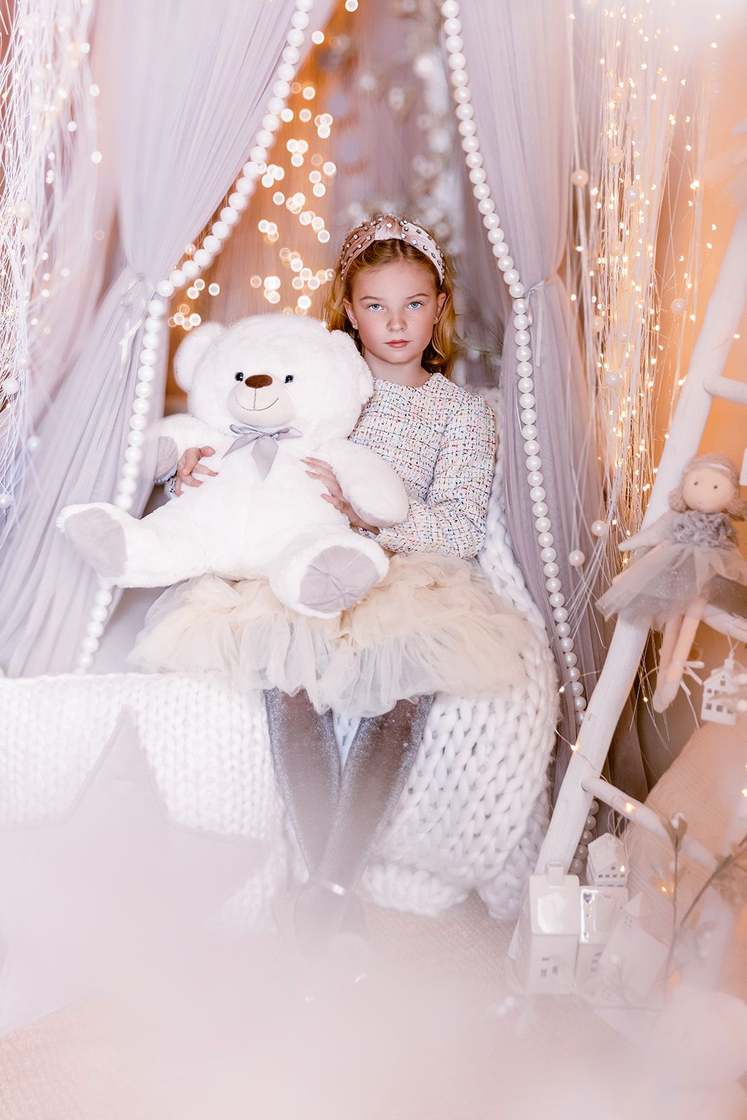 Young girl in white dress holds large white teddy bear on decorated bed with fairy lights and pearl garlands.