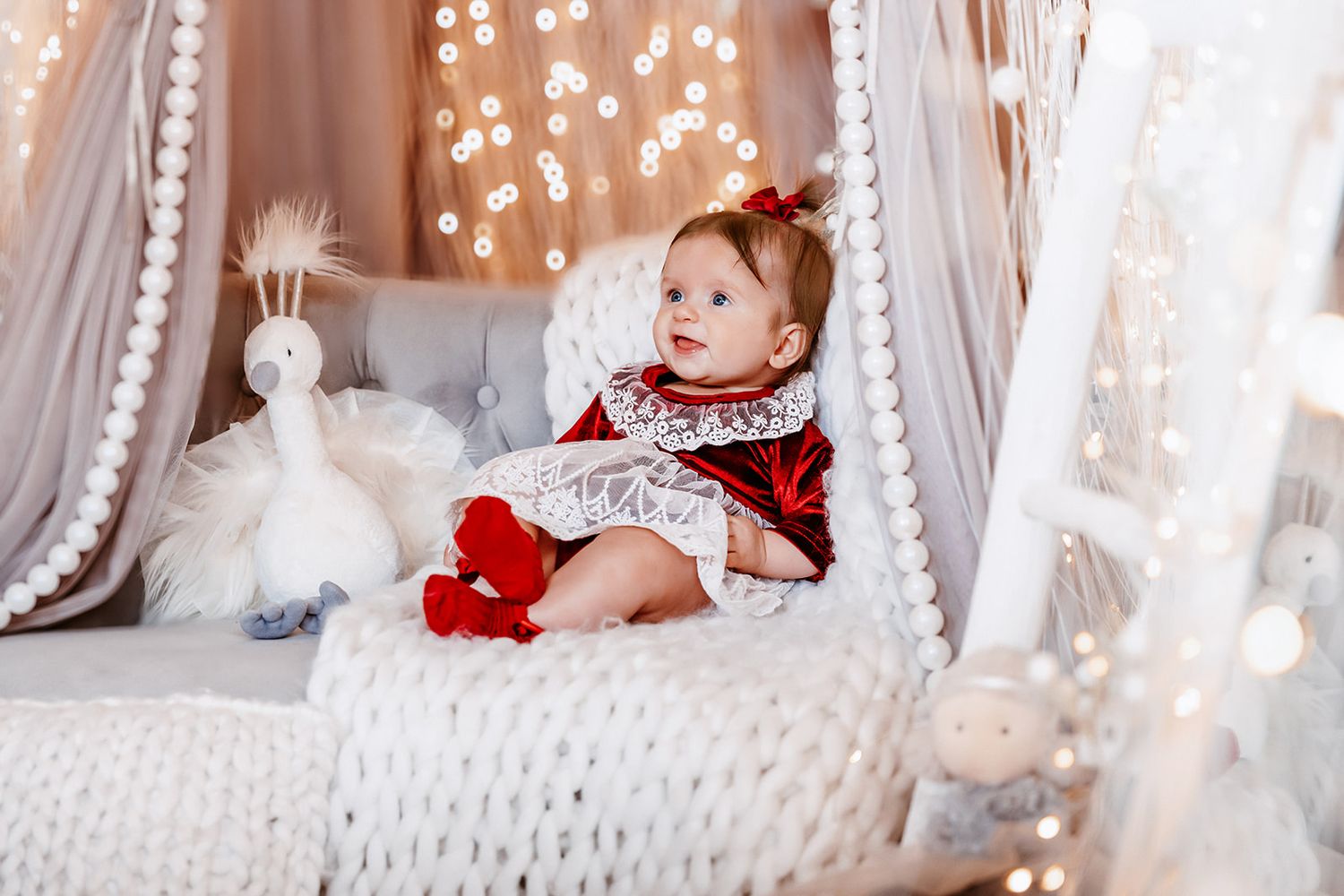 Baby girl in red velvet dress sits in white canopy bed with bokeh lights and swan toy.