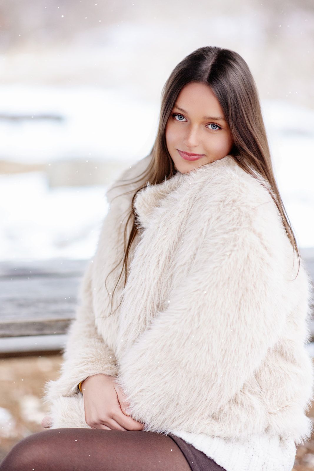 A beautiful teenage girl with blue eyes wearing a cream faux fur coat poses gracefully in a snowy winter setting.