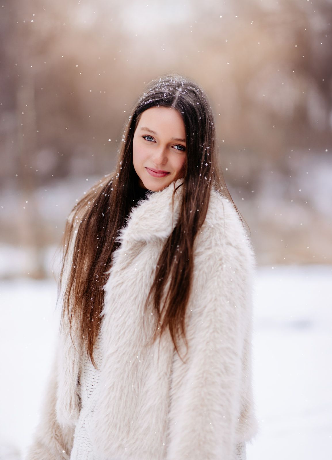 Young woman with long brown hair wearing a white fluffy coat standing outdoors in a snowy winter landscape.