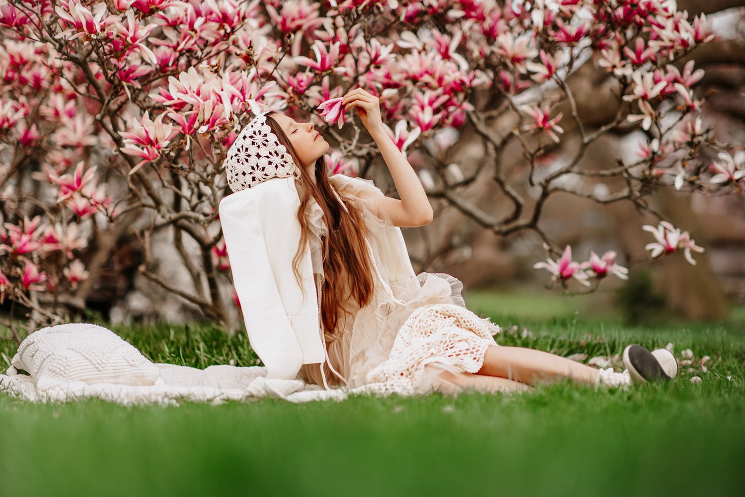 A girl in a white lace dress sits on a blanket beneath a blooming pink magnolia tree in spring.
